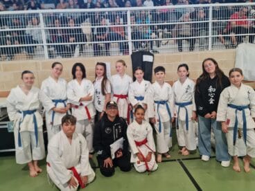 Grupo de jóvenes estudiantes de karate Washinkai y sus instructores posando en un gimnasio con público al fondo.