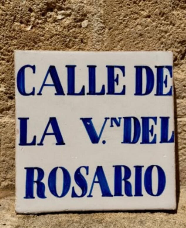 Abbildung: Altes Keramikschild mit blauen Buchstaben und der Aufschrift "CALLE DE LA V.NA DEL ROSARIO" auf weißem Hintergrund.