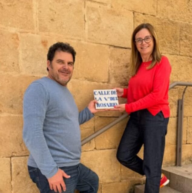 Bild: Ein Mann und eine Frau halten vor einer Steinmauer eine alte Fliese mit der Aufschrift „CALLE DE LA V. DEL ROSARIO“.