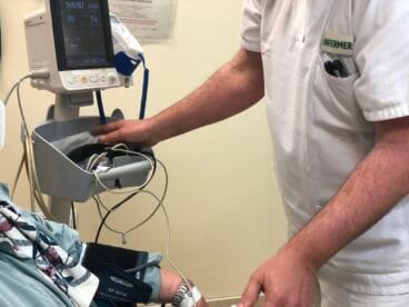 Un enfermero con uniforme blanco y el texto "INFERMER" toma la presión arterial a un paciente, cuyo brazo tiene un manguito conectado a un monitor de signos vitales.