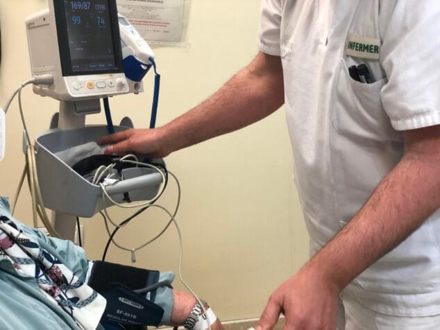 Un enfermero con uniforme blanco y el texto "INFERMER" toma la presión arterial a un paciente, cuyo brazo tiene un manguito conectado a un monitor de signos vitales.