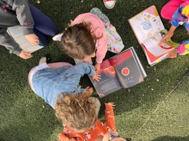 Vista cenital de cuatro niños pequeños tumbados en el césped leyendo libros juntos.