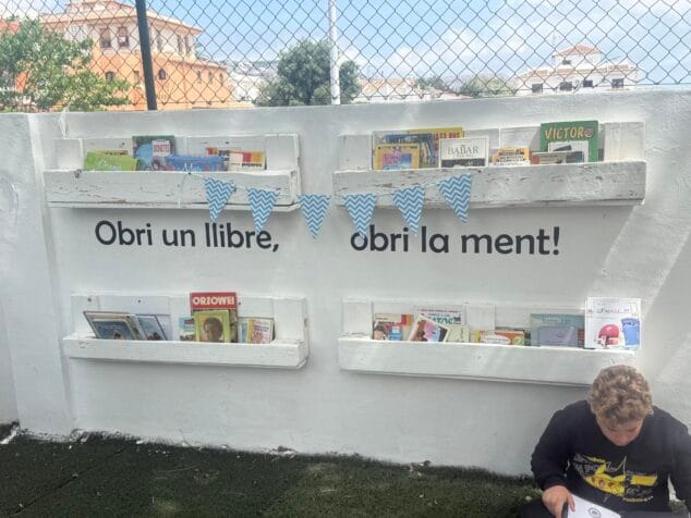 Imagen: Fotografía de un rincón de intercambio de libros infantil al aire libre, con estanterías hechas de palés blancos llenas de cuentos y el texto 'Obri un llibre, obri la ment!' en la pared. Un niño lee sentado en el suelo.