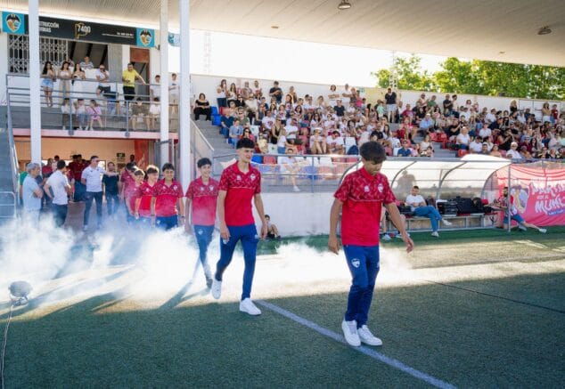 Imagen: Varios jugadores jóvenes del CD Jávea con equipación roja y azul caminan sobre el césped del estadio saliendo de una nube de humo blanco, mientras el público les observa desde la grada durante la fiesta de final de temporada.