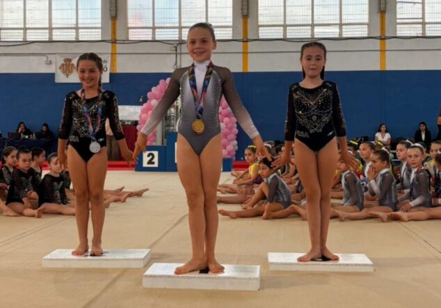 Tres jóvenes gimnastas en un podio con medallas dentro de un pabellón deportivo.