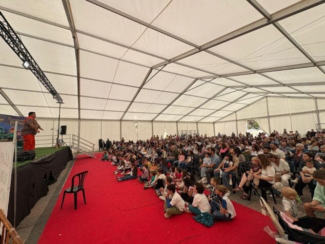 Un hombre actúa sobre un escenario frente a una gran audiencia de niños y adultos bajo una carpa blanca.