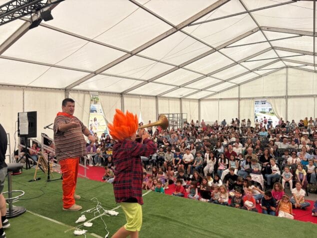 Paco Cholbi en el escenario con un niño con peluca naranja tocando una trompeta, ante un público numeroso bajo una carpa blanca.