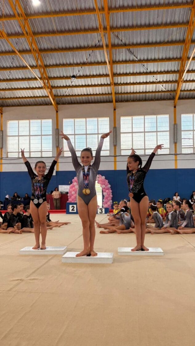 Imagen: Tres jóvenes gimnastas del CD Montgo en lo alto de un podio, con los brazos levantados y medallas al cuello, en un pabellón deportivo.