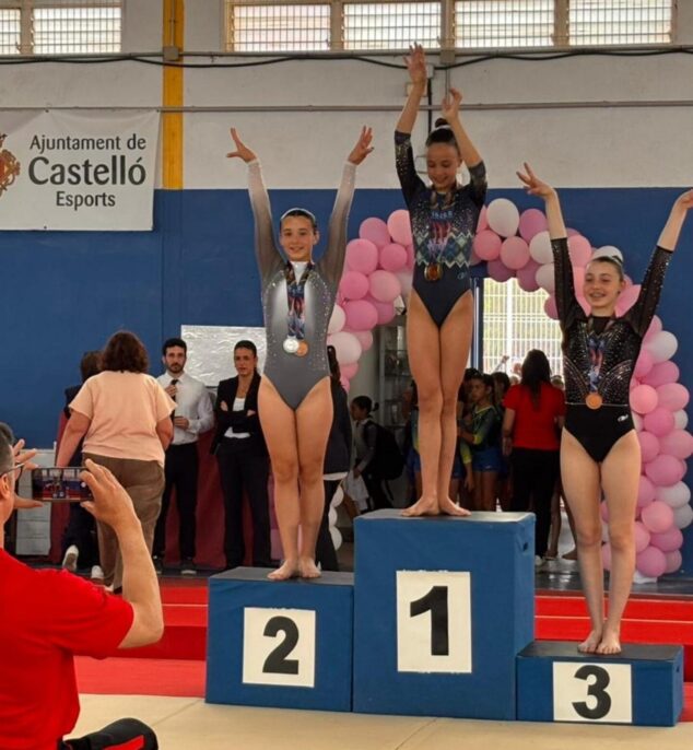 Tres gimnastas jóvenes en un podio con medallas y los brazos levantados, celebrando en un pabellón deportivo.