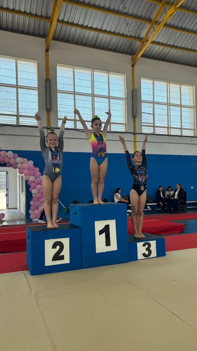 Imagen: Tres gimnastas del Club Deportivo Montgo levantan los brazos con sus medallas en un podio.