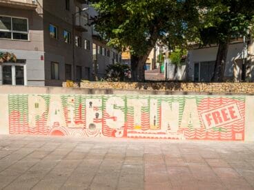Mural de esgrafiado en una pared de piedra con el texto "PALESTINA FREE" y elementos simbólicos palestinos en tonos rojos y verdes.