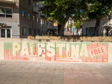 Mural de esgrafiado en una pared de piedra con el texto "PALESTINA FREE" y elementos simbólicos palestinos en tonos rojos y verdes.