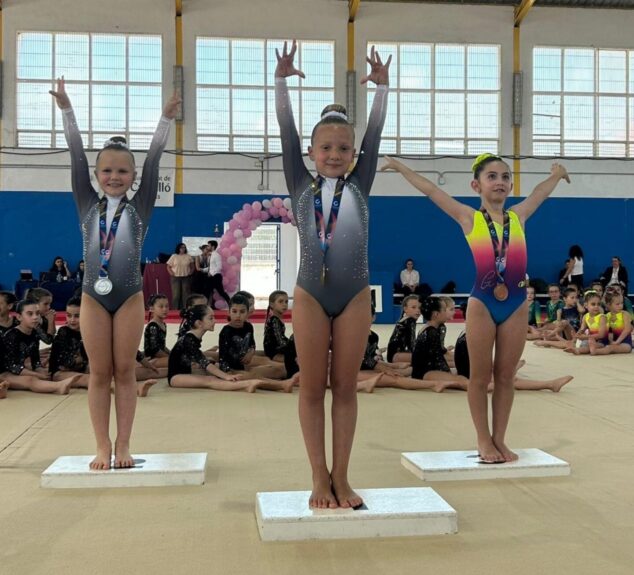 Tres jóvenes gimnastas del Club Montgo Xàbia en el podio con medallas y brazos levantados en un pabellón deportivo.