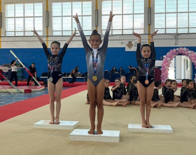 Tres jóvenes gimnastas en un podio deportivo con medallas al cuello y los brazos levantados en señal de victoria.
