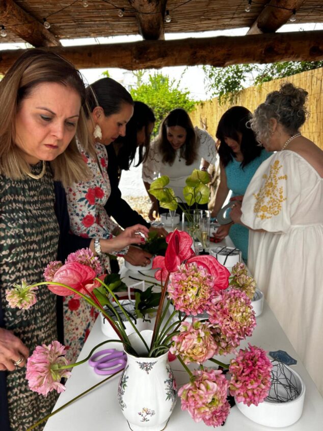 Imagen: Varias mujeres participan en un taller de arreglos florales con flores rosas y verdes sobre una mesa blanca al aire libre.