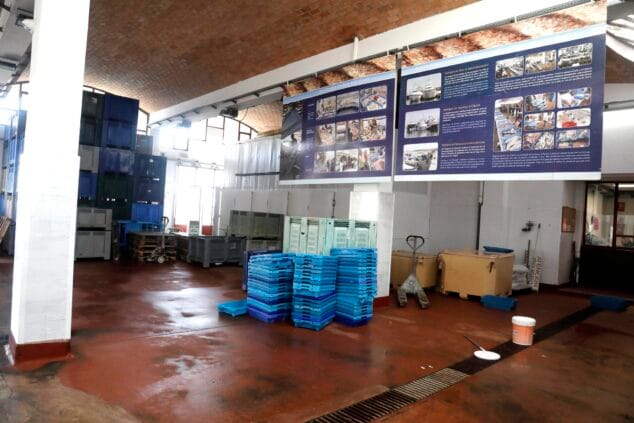 Interior de una lonja de pescado con pilas de cajas de plástico azules y grises y paneles informativos en la pared de fondo.