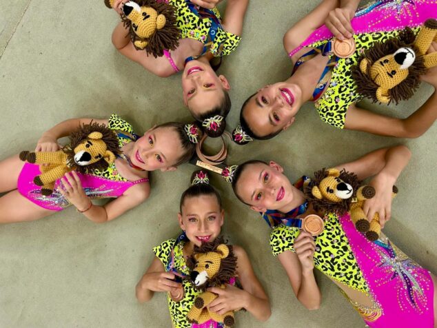 Imagen: Cinco gimnastas del conjunto alevín de GR Xàbia tumbadas en círculo, sonriendo y mostrando sus medallas de bronce y peluches de león.