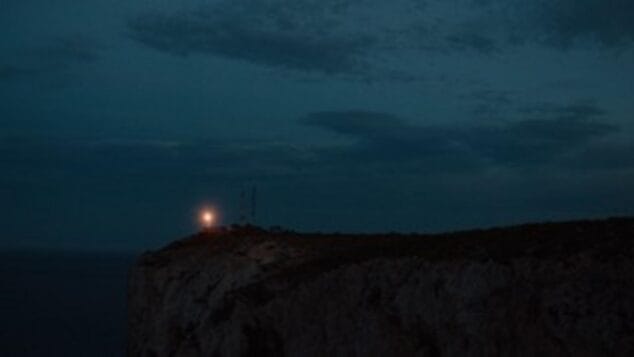 Imagen: Fotograma oscuro y atmosférico de la película 'AILEEN' que muestra una luz sobre un acantilado junto al mar.