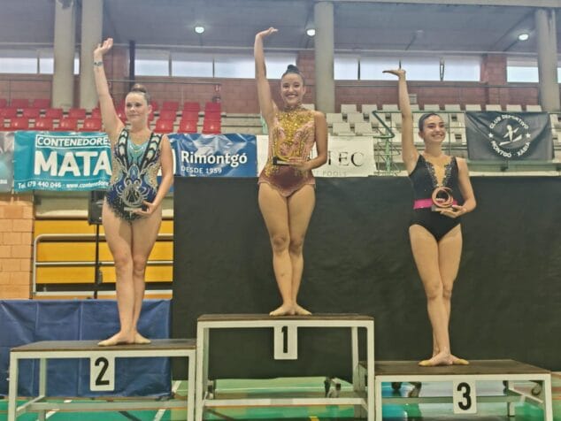 Imagen: Tres gimnastas rítmicas con trofeos saludando desde el podio en un pabellón deportivo.