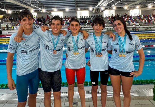 Imagen: Cinco nadadores del Club Natació Dénia posan con medallas al borde de una piscina cubierta tras una competición.