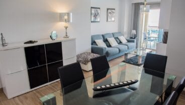 alón-comedor de concepto abierto con mesa de cristal, sillas negras y sofá azul. El suelo de baldosas y un aparador blanco moderno complementan el espacio, con acceso a la terraza