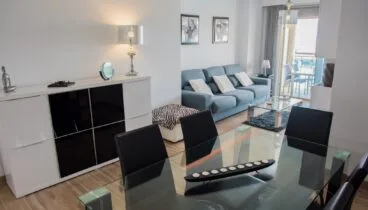alón-comedor de concepto abierto con mesa de cristal, sillas negras y sofá azul. El suelo de baldosas y un aparador blanco moderno complementan el espacio, con acceso a la terraza