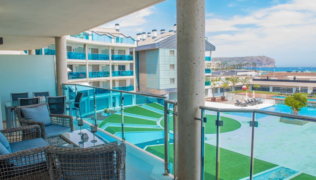 Amplia terraza privada con mobiliario de exterior de ratán y vistas directas a las piscinas comunitarias y al mar. Barandilla de cristal y suelo de gres claro