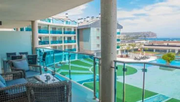 Amplia terraza privada con mobiliario de exterior de ratán y vistas directas a las piscinas comunitarias y al mar. Barandilla de cristal y suelo de gres claro