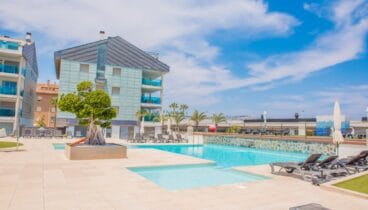 Amplia zona de piscina comunitaria con dos piscinas, pavimentación clara y numerosas tumbonas. Edificios residenciales modernos con balcones de cristal azulado enmarcan este espacio soleado