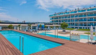 Amplia zona de piscina comunitaria con una piscina de azul brillante y una tarima de madera cálida. El moderno edificio de apartamentos, con balcones de cristal azul, domina este espacio exterior sereno