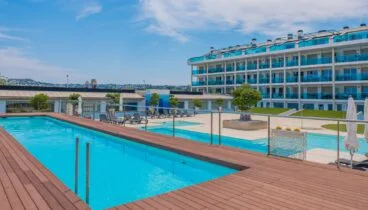 Amplia zona de piscina comunitaria con una piscina de azul brillante y una tarima de madera cálida. El moderno edificio de apartamentos, con balcones de cristal azul, domina este espacio exterior sereno