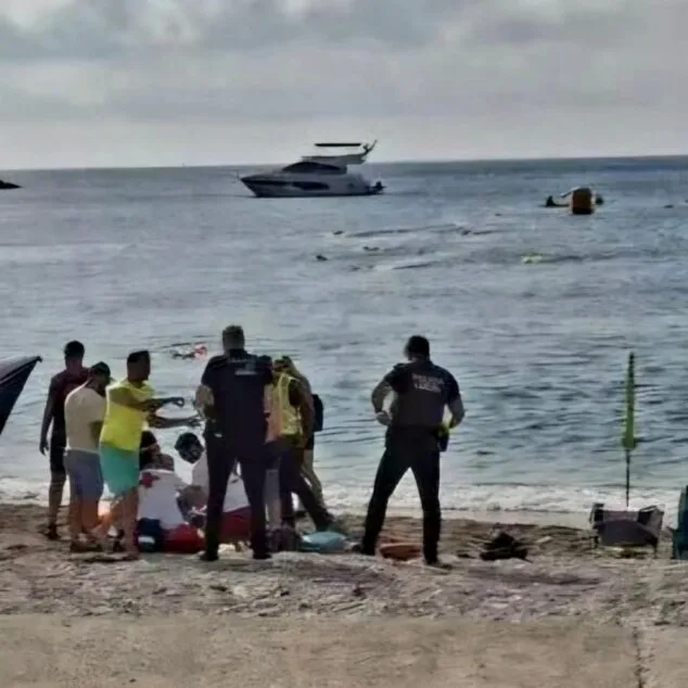 Varios agentes de policía y personal de emergencias asisten a una persona tendida en la arena de la playa del Arenal, con el mar y un barco al fondo.