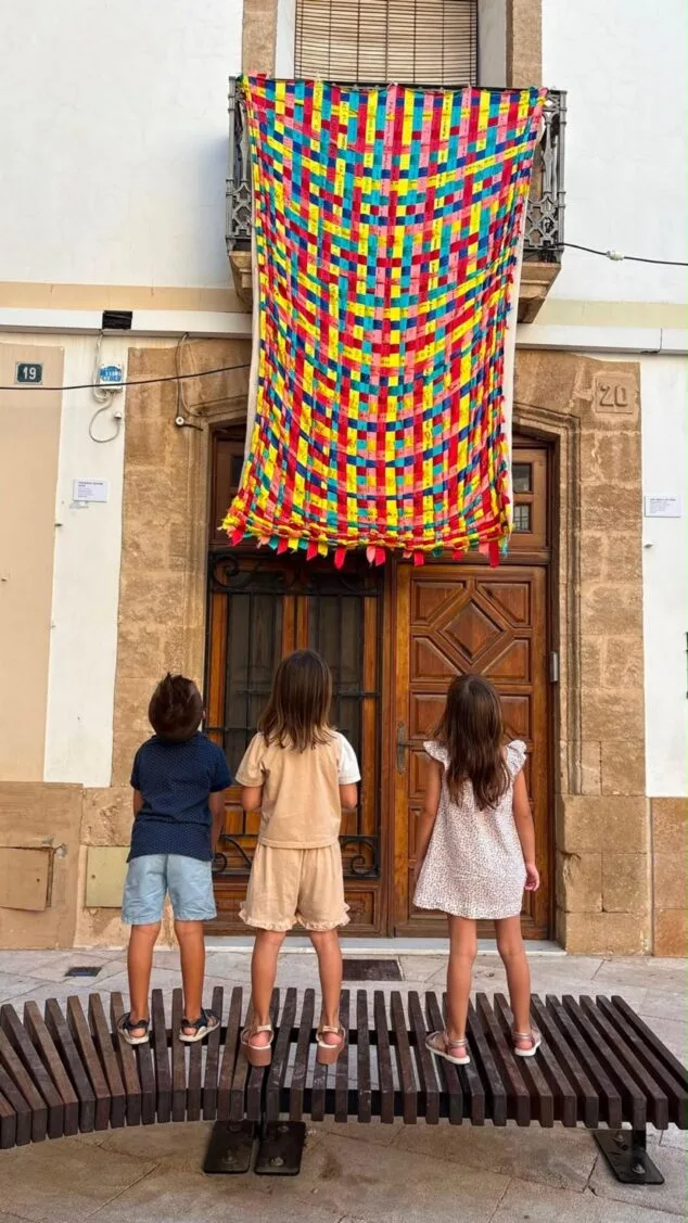 Imagen: Tres niños de espaldas miran una gran tela colorida hecha de cintas entrelazadas que cuelga de un balcón.