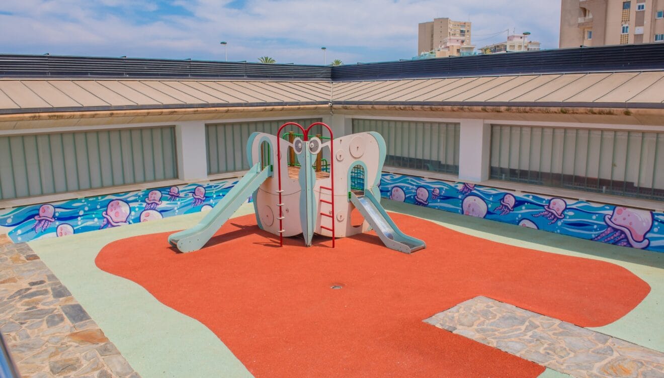 Colorida zona de juegos infantiles con estructura central de toboganes sobre superficie de caucho rojo y verde. Un vibrante mural de medusas decora la pared, bajo paneles de vidrio esmerilado