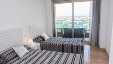 Dormitorio luminoso con dos camas individuales, ropa de cama a rayas y mesita de noche. Puertas correderas dan a un balcón con asientos exteriores y vistas parciales al mar. Suelo pulido