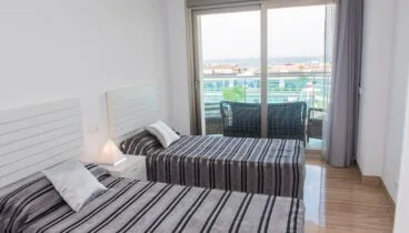 Dormitorio luminoso con dos camas individuales, ropa de cama a rayas y mesita de noche. Puertas correderas dan a un balcón con asientos exteriores y vistas parciales al mar. Suelo pulido