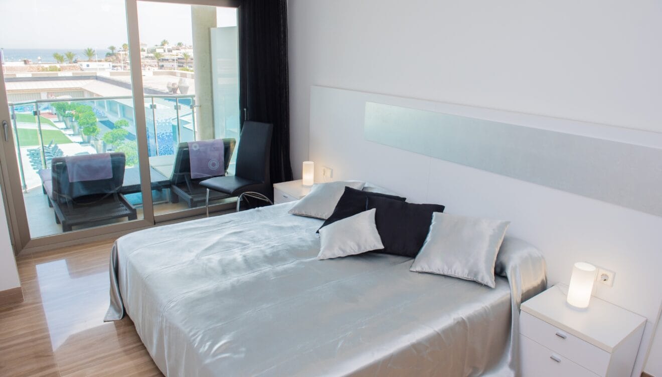 Dormitorio principal moderno con cama grande, ropa de cama satinada plateada, suelos de gres brillante y puertas correderas a la terraza con vistas al mar y piscina