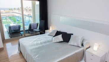 Dormitorio principal moderno con cama grande, ropa de cama satinada plateada, suelos de gres brillante y puertas correderas a la terraza con vistas al mar y piscina