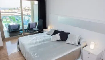 Dormitorio principal moderno con cama grande, ropa de cama satinada plateada, suelos de gres brillante y puertas correderas a la terraza con vistas al mar y piscina