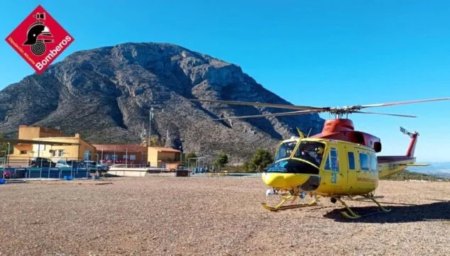 Helicóptero de rescate amarillo y rojo de los Bomberos de la Diputación de Alicante estacionado con la montaña del Montgó al fondo.