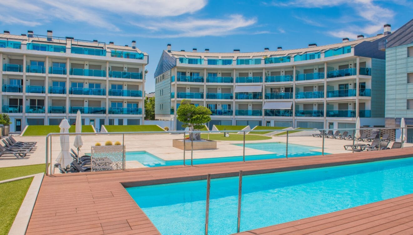 La zona de piscina comunitaria presenta dos piscinas de agua azul cristalina, rodeadas de tarima de madera, tumbonas y césped artificial, frente a edificios de apartamentos modernos