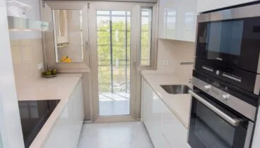 Moderna cocina tipo galera con elegantes muebles blancos, encimeras claras y electrodomésticos Siemens integrados. La puerta doble acristalada proporciona luz natural y acceso a un balcón