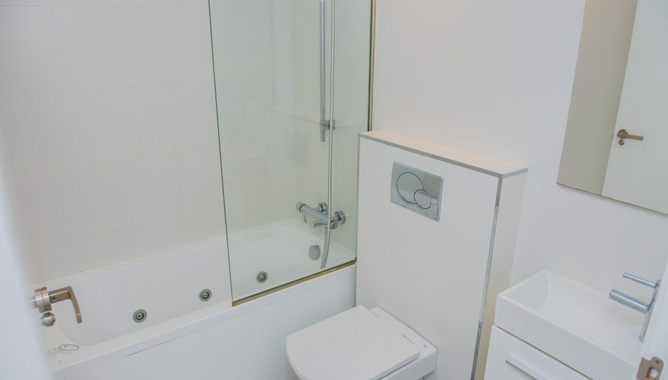 Moderno cuarto de baño blanco con bañera de hidromasaje, mampara de cristal, inodoro suspendido y lavabo rectangular. Azulejos blancos y grifería cromada