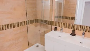 Moderno cuarto de baño con ducha a ras de suelo y mampara de cristal. Destaca el lavabo doble y los azulejos beige con franja decorativa de mosaico