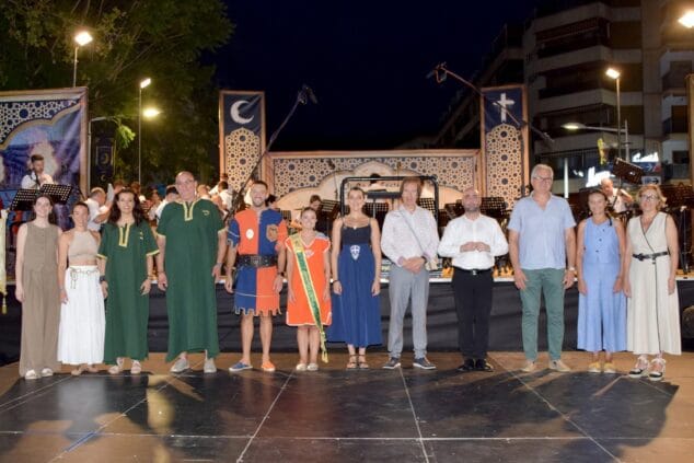 Imagen: Grupo de personas, incluyendo cargos festeros con trajes tradicionales, posando en un escenario al aire libre por la noche tras un concierto.