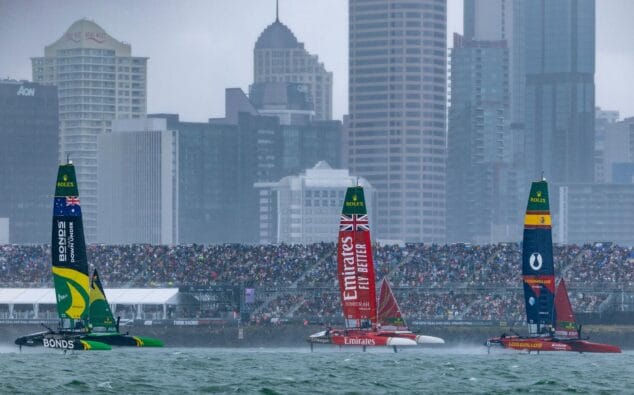 Imagen: Catamaranes F50 de España, Gran Bretaña y Australia compitiendo en una regata de SailGP bajo la lluvia y con edificios de fondo.