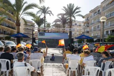 Personas viendo una carrera de vela en una pantalla gigante al aire libre en Xàbia, con banderas de España.