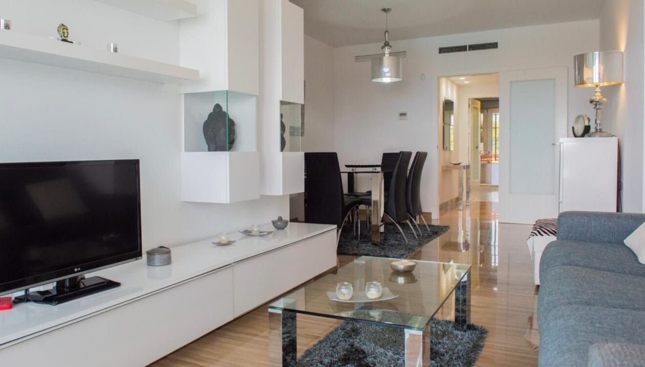 Salón-comedor moderno con mueble modular blanco, mesa de centro de cristal sobre alfombra oscura y conjunto de comedor con sillas negras. Suelos pulidos y aire acondicionado integrado