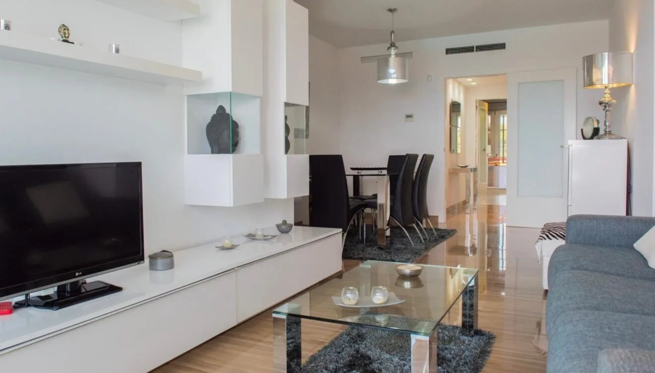 Salón-comedor moderno con mueble modular blanco, mesa de centro de cristal sobre alfombra oscura y conjunto de comedor con sillas negras. Suelos pulidos y aire acondicionado integrado