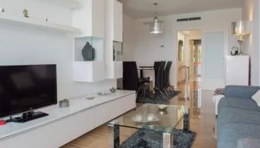 Salón-comedor moderno con mueble modular blanco, mesa de centro de cristal sobre alfombra oscura y conjunto de comedor con sillas negras. Suelos pulidos y aire acondicionado integrado